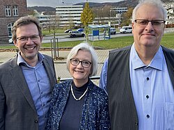 Drei lächelnde Erwachsene, zwei Männer und eine Frau, im Freien.