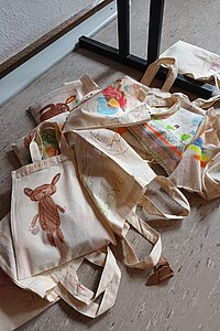 Mehrere Stofftaschen mit Kinderzeichnungen liegen auf dem Boden.