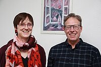 Lächelnde Frau und Mann stehen vor einem gerahmten Gemälde.